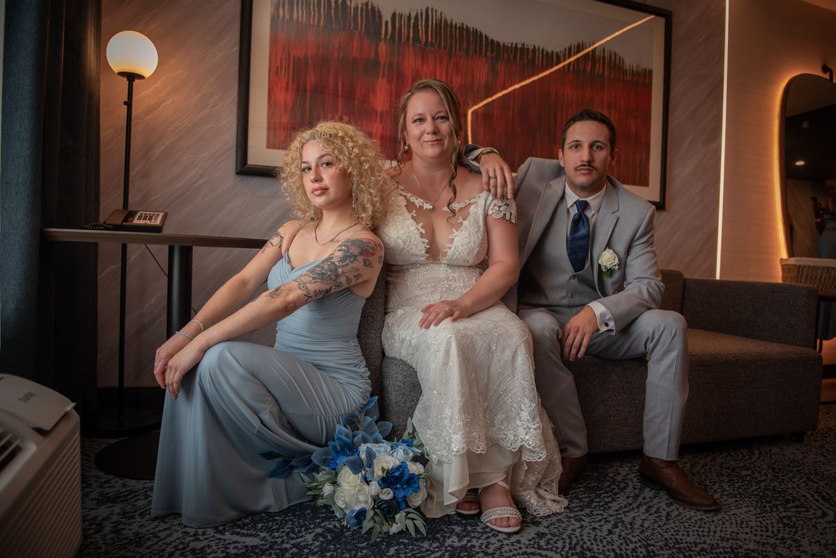 A bride in white lace sits between a friend in blue and the groom in gray, surrounded by soft lighting and modern decor.