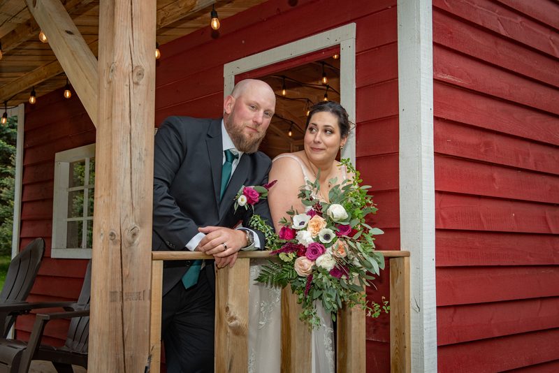 Anastasia and Adam stand on a rustic wooden porch of a red-sided cabin, gazing at each other with smiles.