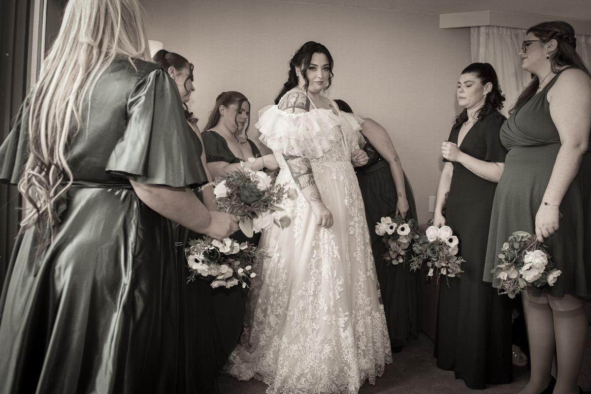 A bride with tattoos and her bridesmaids in green dresses hold floral bouquets, preparing for the ceremony.