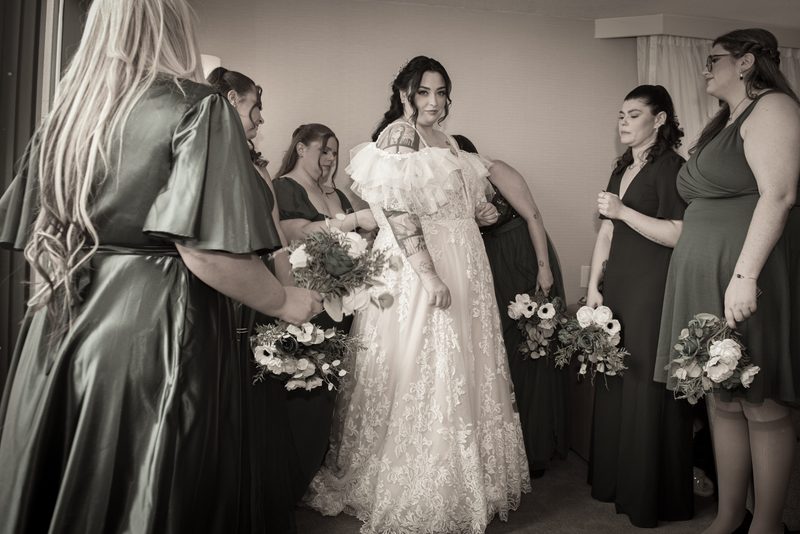 A bride with tattoos and her bridesmaids in green dresses hold floral bouquets, preparing for the ceremony.