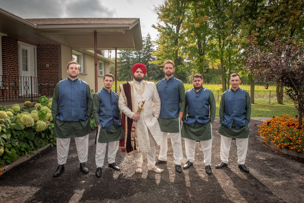 A groom in traditional attire stands with four groomsmen posing outdoors in front of a house surrounded by autumn foliage.
