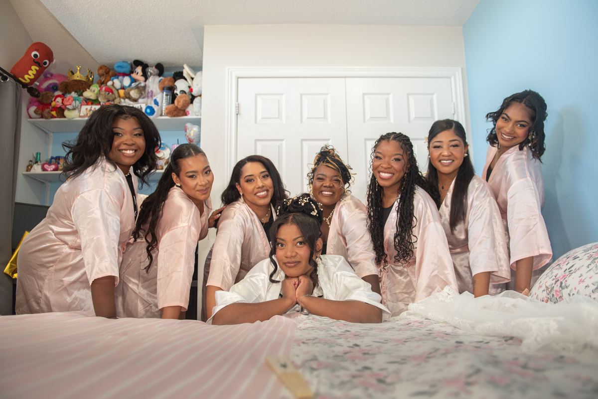 Bride and bridesmaids in pink satin robes smile around bed with floral comforter, room filled with colorful plush toys.