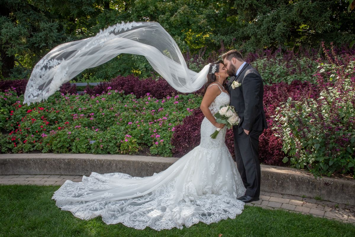 A bride and groom kiss in a sunlit garden with billowing veil and vibrant purple and pink flowers as backdrop.