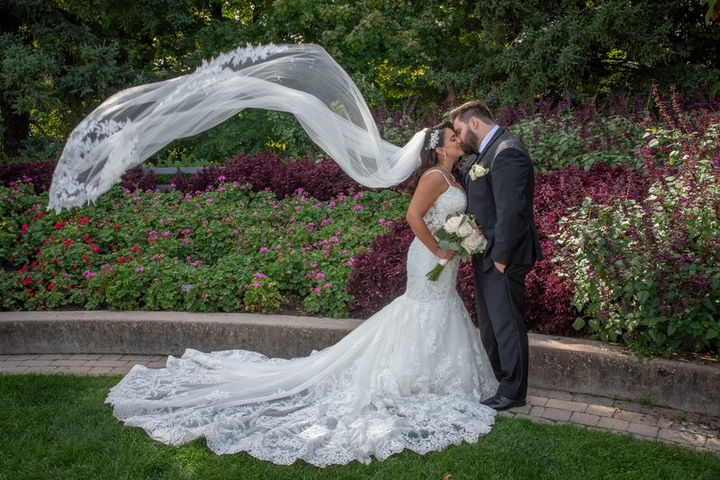 A bride and groom kiss in a sunlit garden with billowing veil and vibrant purple and pink flowers as backdrop.