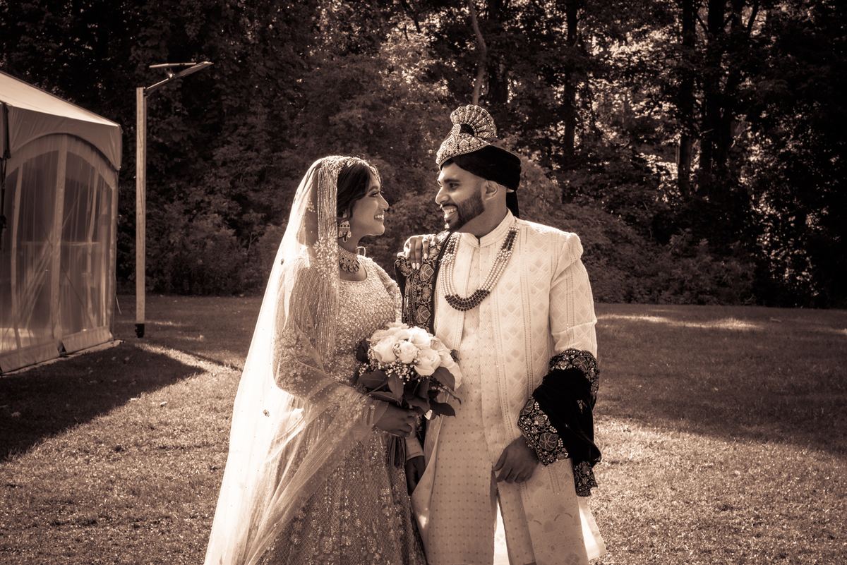 A newlywed couple in traditional attire stands outdoors, smiling at each other in a sun-dappled garden.