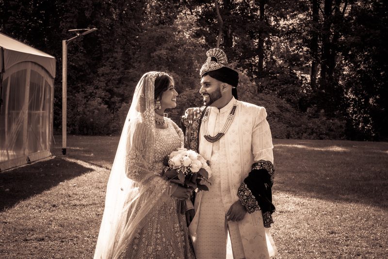 A newlywed couple in traditional attire stands outdoors, smiling at each other in a sun-dappled garden.