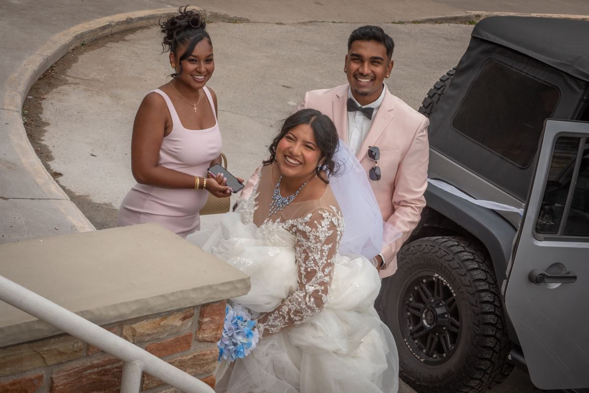 A bride in a white lace gown laughs with her pink-suited groom and two bridesmaids beside a gray Jeep under diffused light.