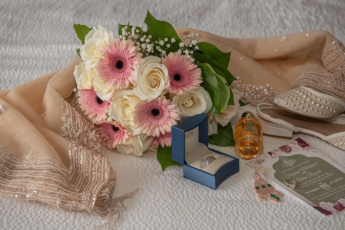 Bride holds pink gerbera and white rose bouquet, blue ring box, perfume bottle, and traditional shawl with footwear.