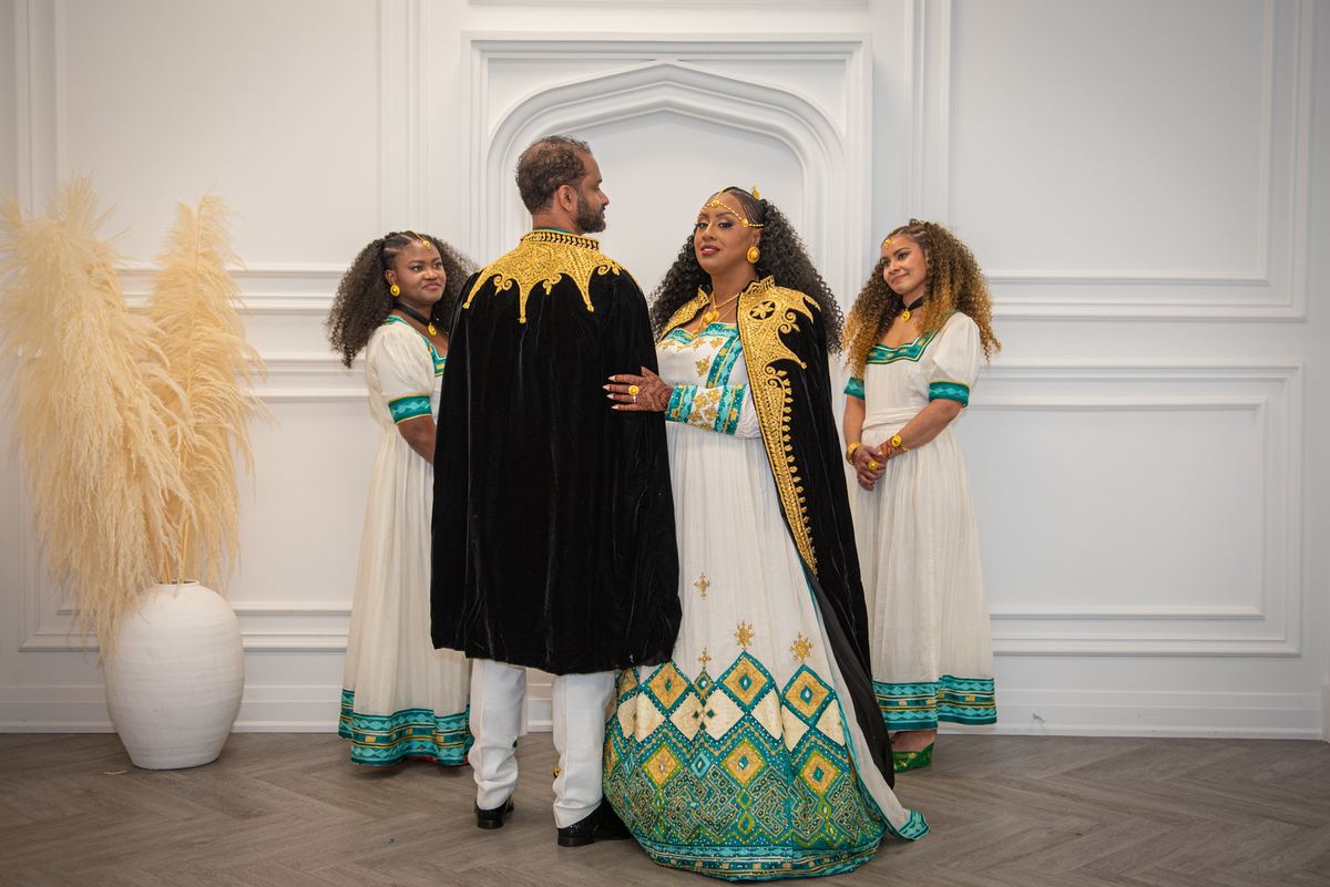 A couple in traditional gold and teal garments stand closely with two women in matching attire near pampas grass.
