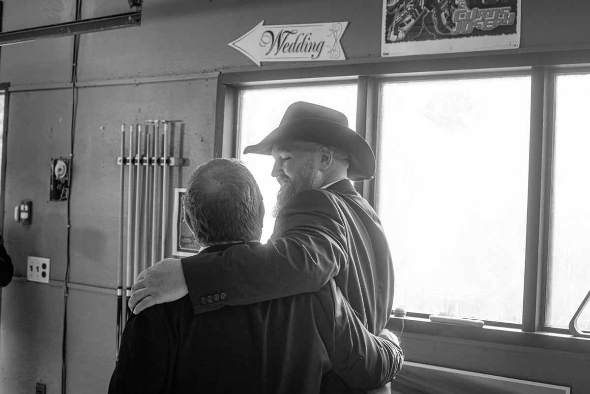 Two men in formal wear embrace near a window, illuminated by natural light, creating an affectionate scene during a wedding.