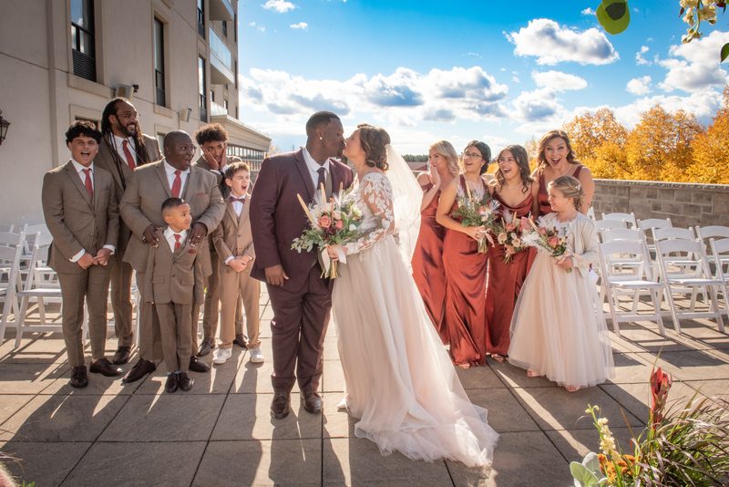 Jennifer and Kevin share a tender kiss at their outdoor ceremony, surrounded by their joyful wedding party.