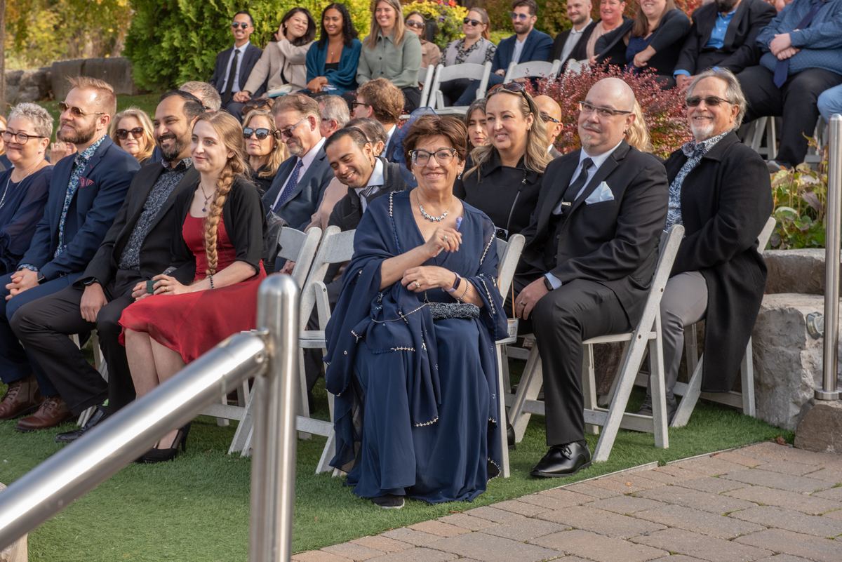 Guests in formal attire sit quietly in white folding chairs, smiling at an unseen focal point during a ceremony.