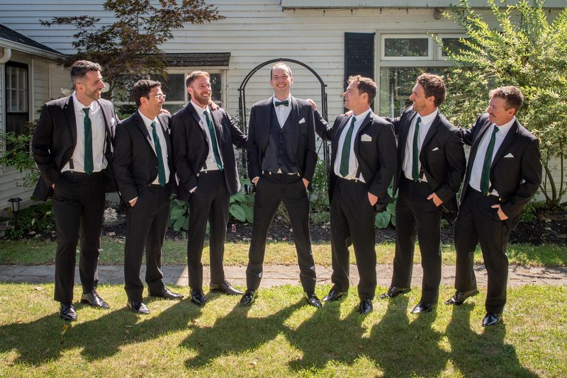 Groomsmen in black suits and green ties laugh together outdoors on a sunny day, casting long shadows.