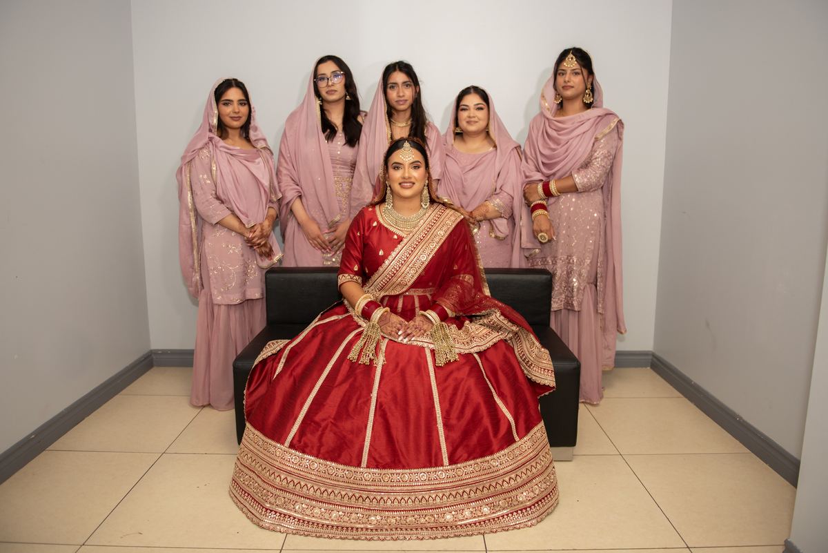A bride in a red and gold lehenga sits on a black couch with five bridesmaids in pink traditional attire behind her.