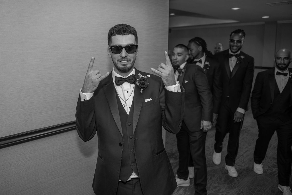 A groom in a tuxedo makes a metal horns gesture, smiling with other formally dressed men behind him in a hotel corridor.