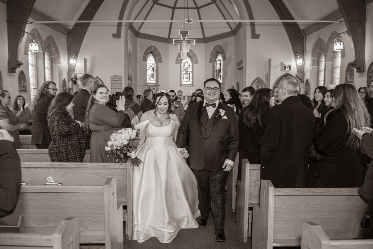 A newlywed couple exits a church aisle surrounded by applauding guests and soft light from stained-glass windows.
