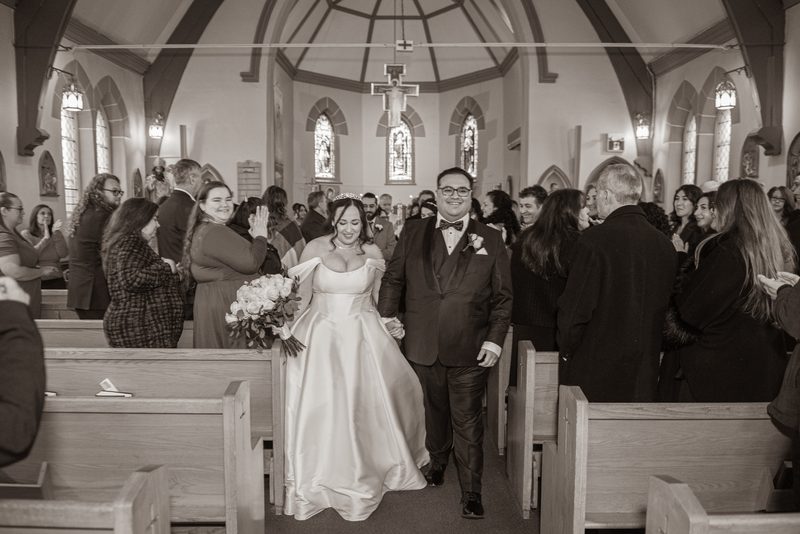 A newlywed couple exits a church aisle surrounded by applauding guests and soft light from stained-glass windows.