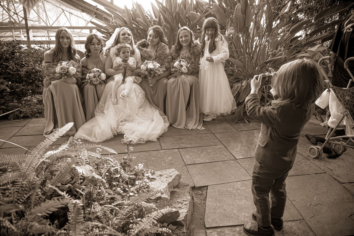 A boy holds a camera while a bride and her bridesmaids, each with a bouquet, sit in a row under a glass greenhouse ceiling.