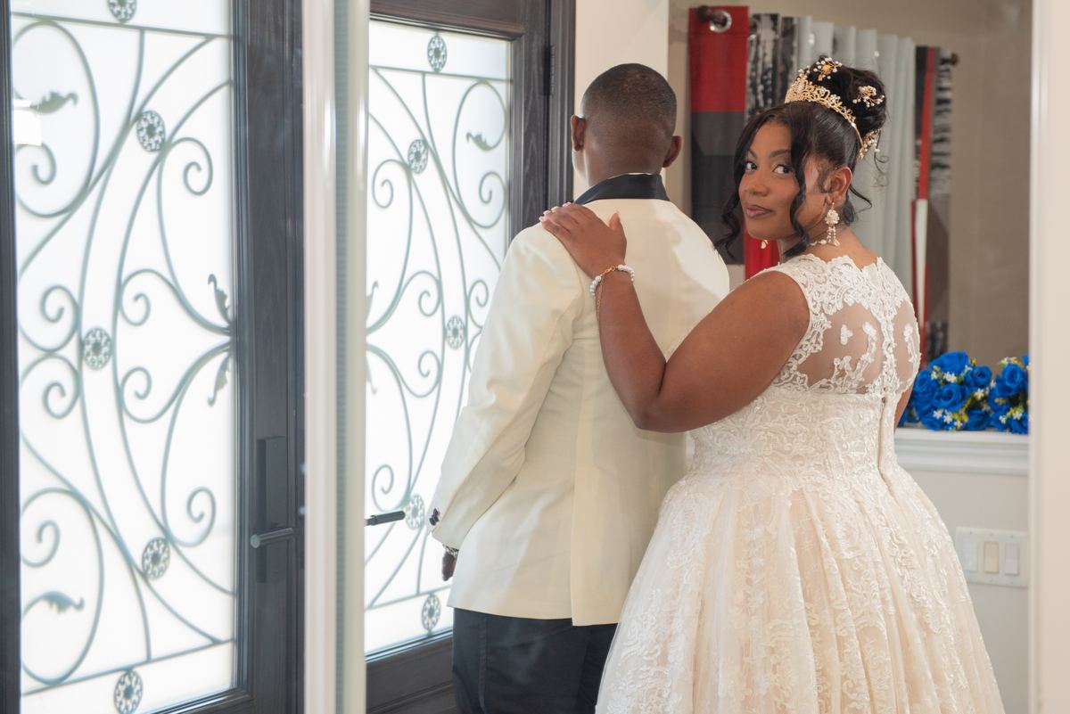 A bride in an elegant lace gown places her hand gently on her groom's shoulder near an ornate glass door.