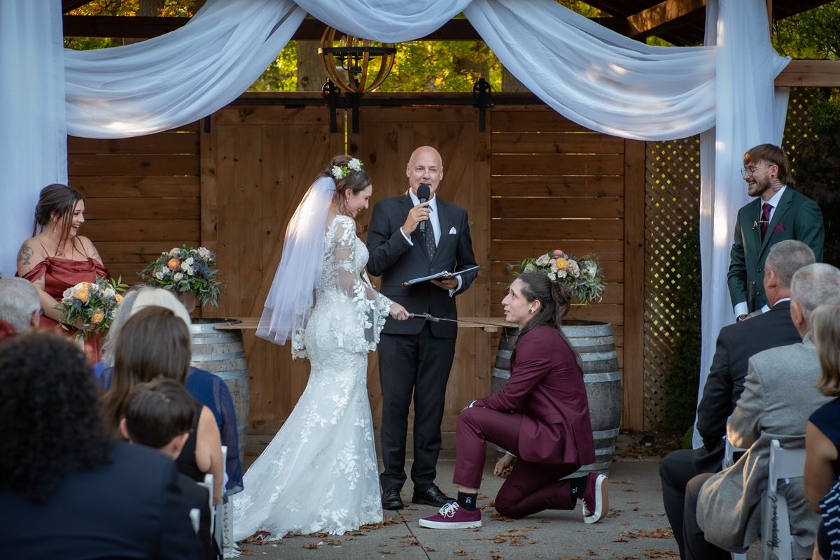 A bride and groom exchange vows under an arbor during an outdoor ceremony with guests in attendance.