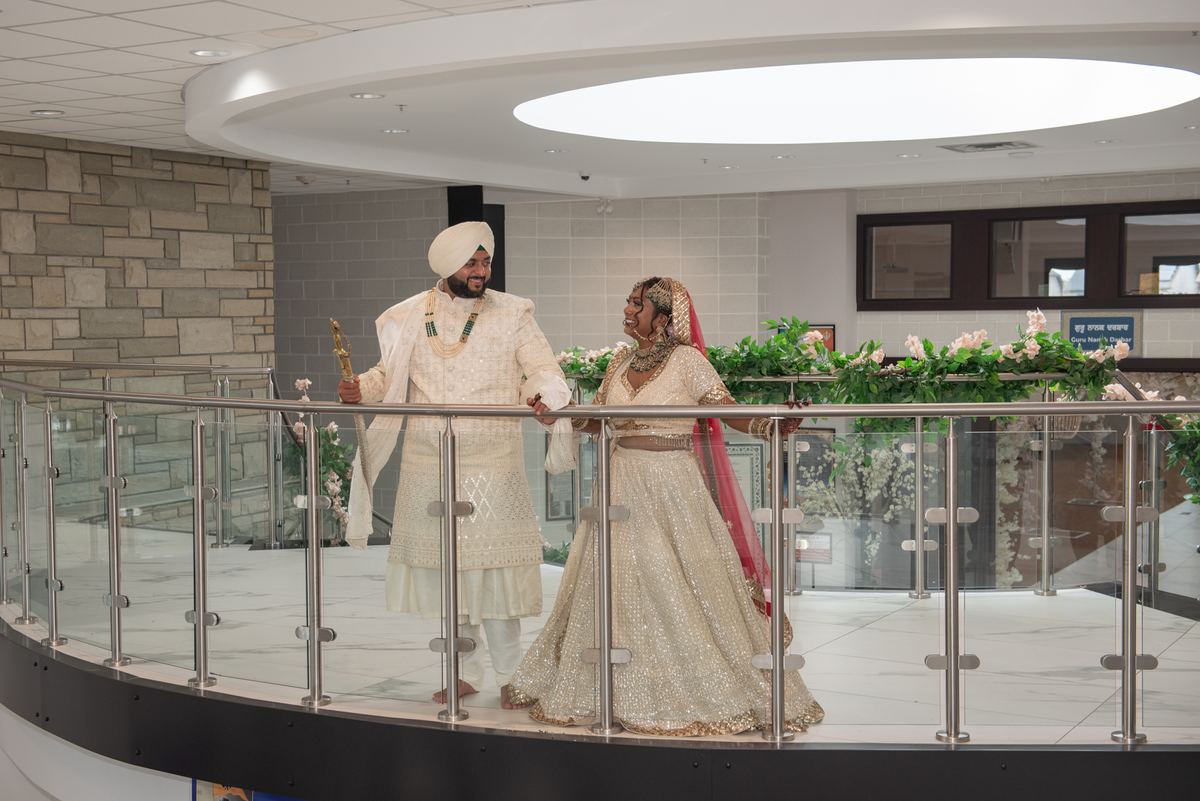 A newlywed Sikh couple in traditional attire walk hand-in-hand along an indoor balcony, surrounded by flowers.