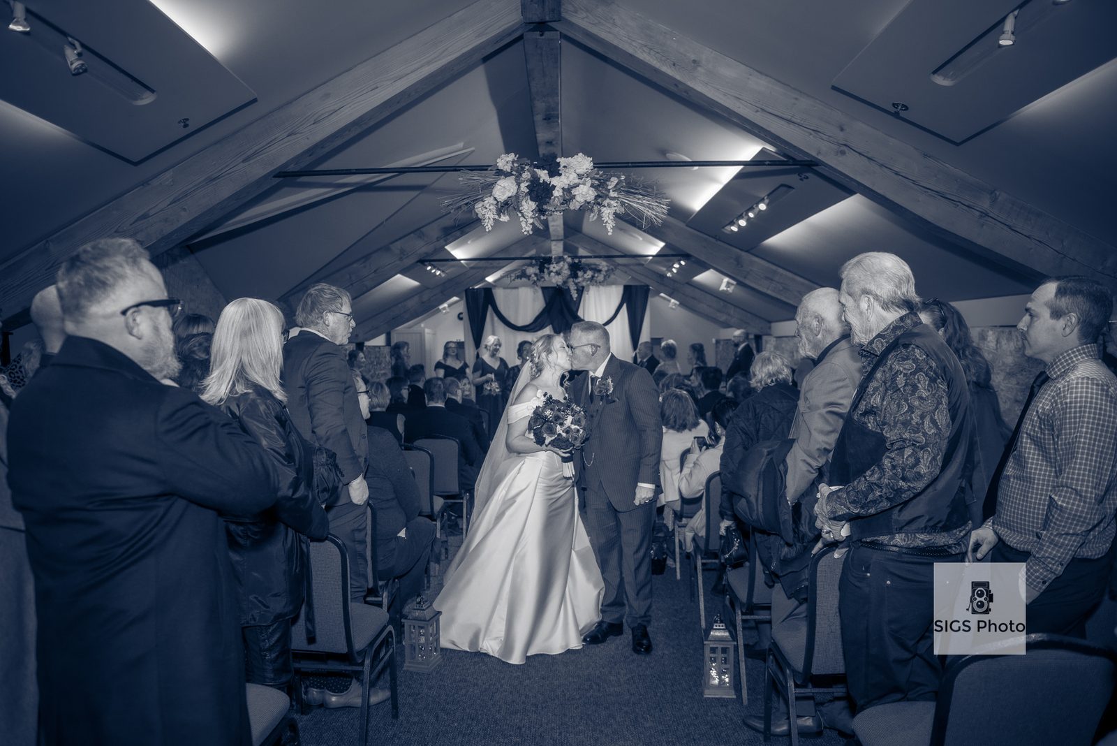 A bride and groom kiss beneath a floral arch in the centre of the aisle as seated guests look on under exposed wooden beams.