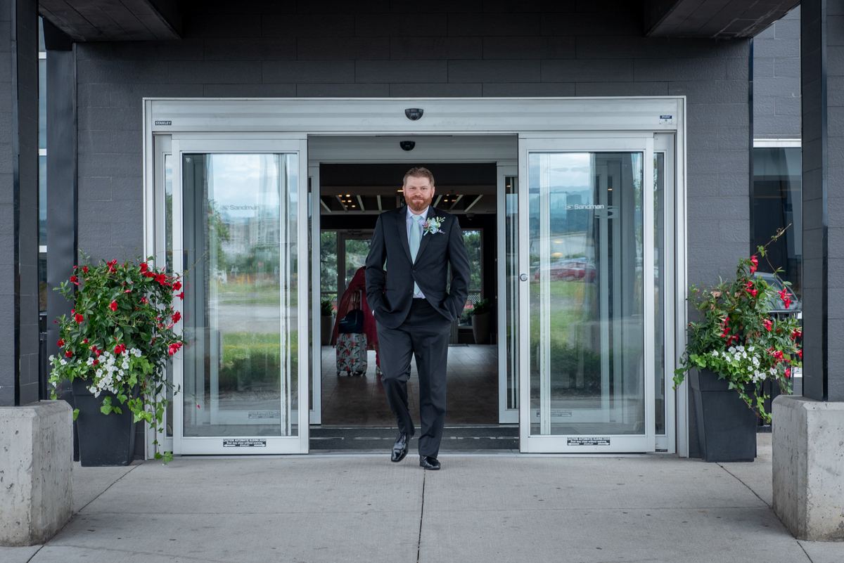 A groom in a dark suit exits glass doors with hands in pockets, passing potted red and white flowering plants.