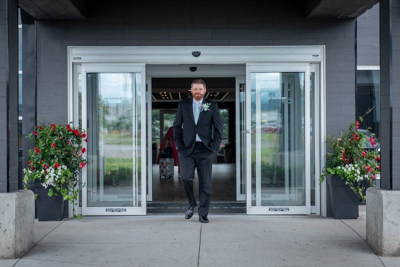 A groom in a dark suit exits glass doors with hands in pockets, passing potted red and white flowering plants.