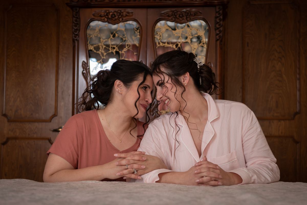 Two women sitting closely on a bed, foreheads touching and smiling warmly in soft, diffused light from patterned windows.