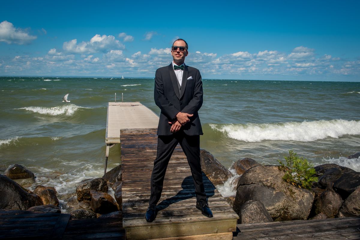 A man in a formal black tuxedo with sunglasses stands confidently on a wooden dock by the sea, waves crashing nearby.