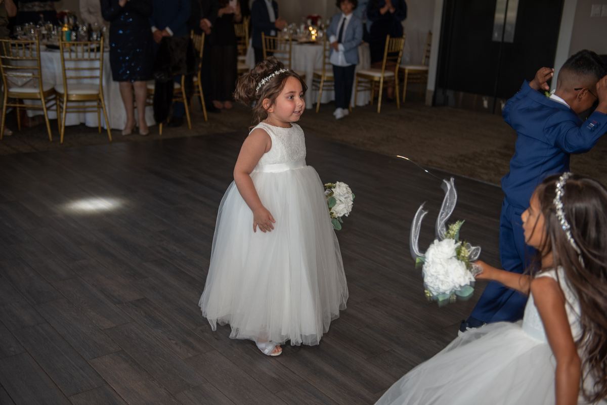 A young flower girl in a white gown walks with a bouquet as another child in blue dances nearby at a wedding.