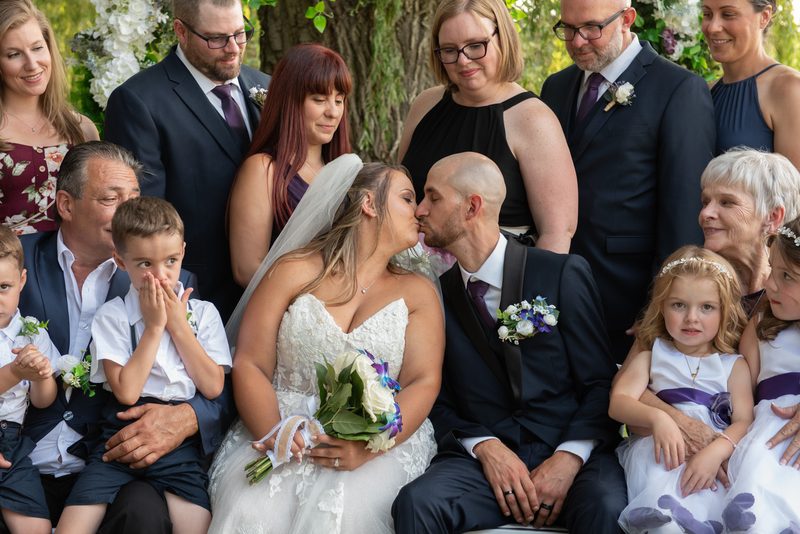 A bride and groom kiss as their wedding party, including children in flower girl dresses, looks on with joy.