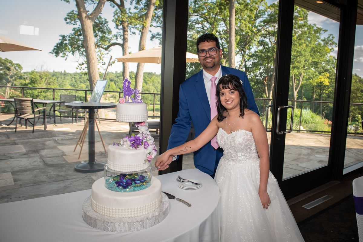 A bride and groom smile as they cut their multi-tiered white cake decorated with purple flowers outdoors on a stone patio.