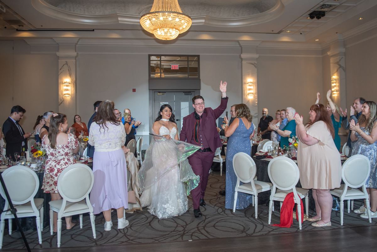 A groom raises one hand as he and his bride dance between tables of clapping guests beneath a large chandelier.