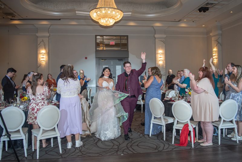 A groom raises one hand as he and his bride dance between tables of clapping guests beneath a large chandelier.