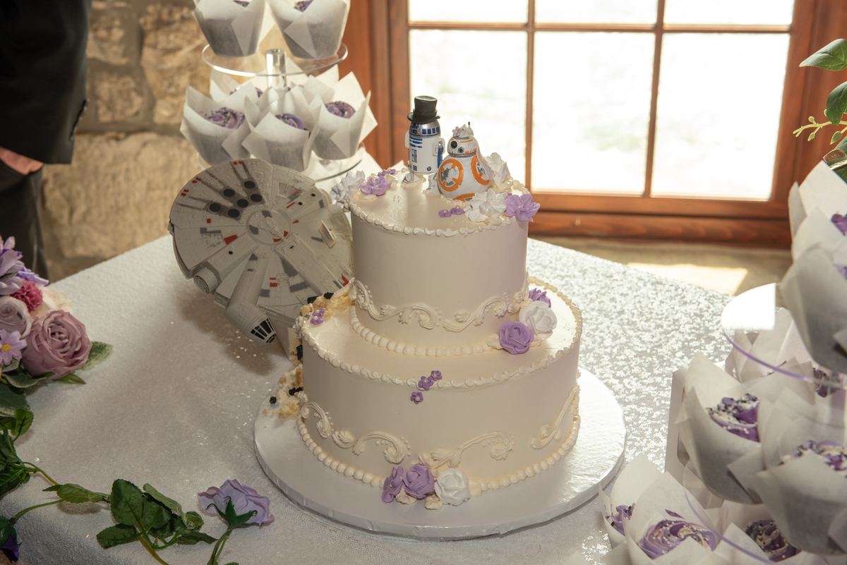 A wedding cake with Star Wars toppers and cupcakes surrounded by flowers, illuminated by natural light.