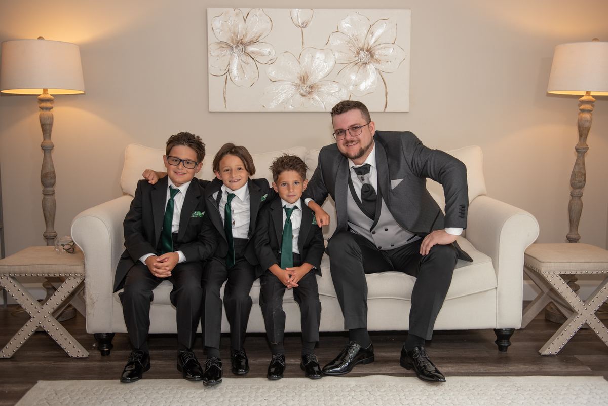 A man and three young boys in matching formal suits with green ties pose on a white sofa in an elegantly decorated room.