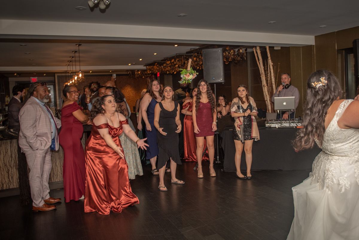 Guests dance and mingle at a DJ booth during a lively wedding reception, with the bride in white visible nearby.