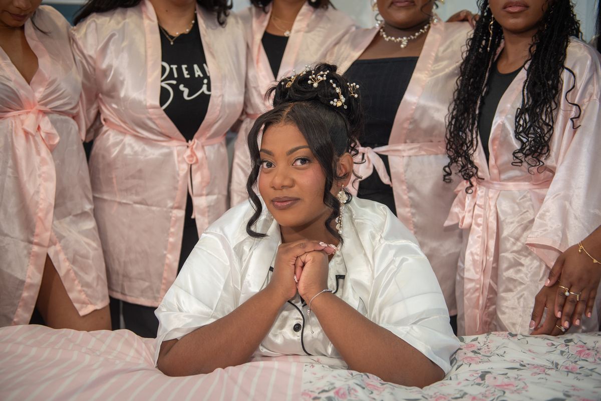 A bride in a white satin robe sits on a bed with her smiling bridesmaids in pink robes behind her, hands clasped.