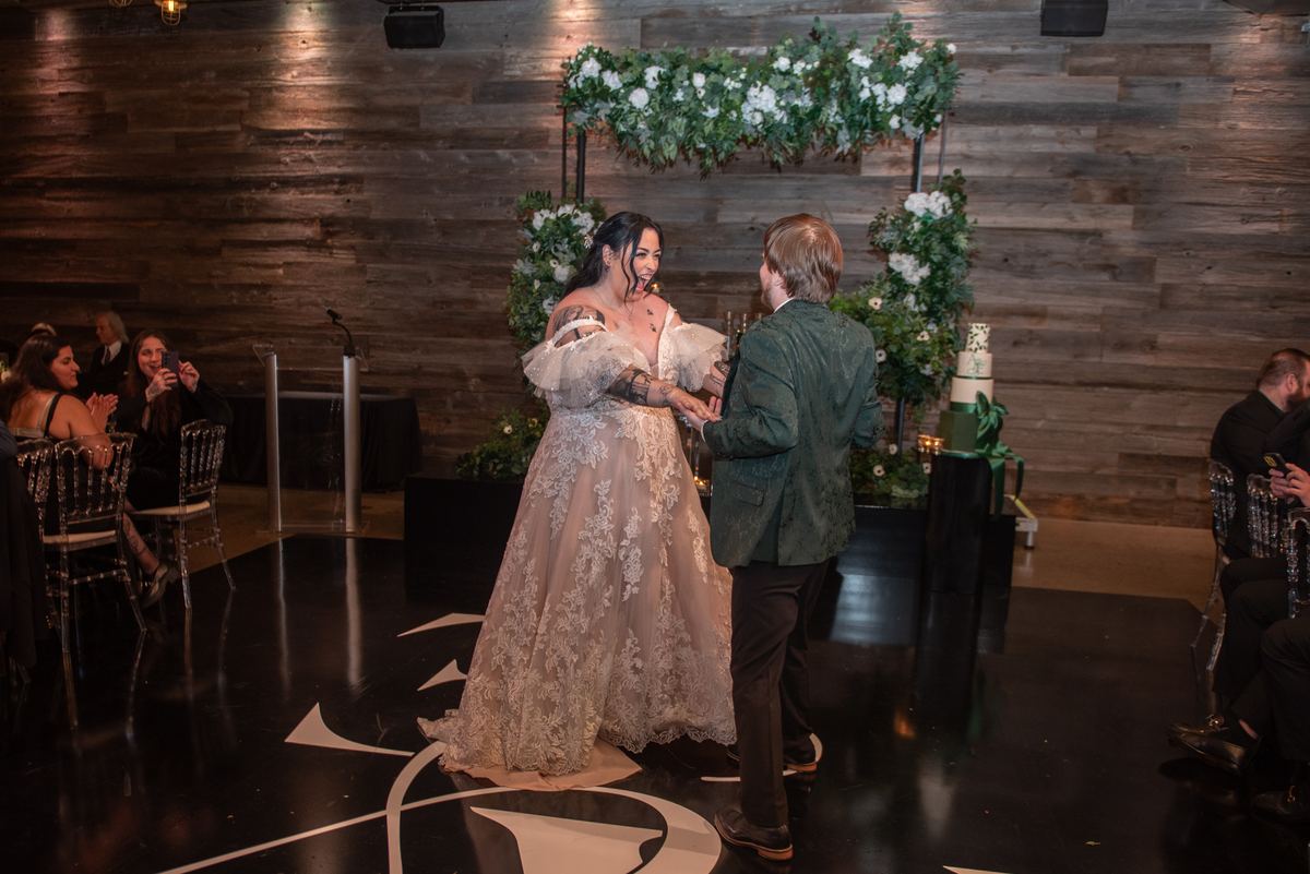 Bride and groom dance intimately on black floor surrounded by guests, rustic wall adorned with greenery and blooms.