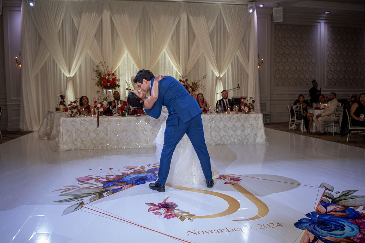 A bride and groom dance intimately on a floral-decorated floor, the groom kissing her forehead as guests watch.