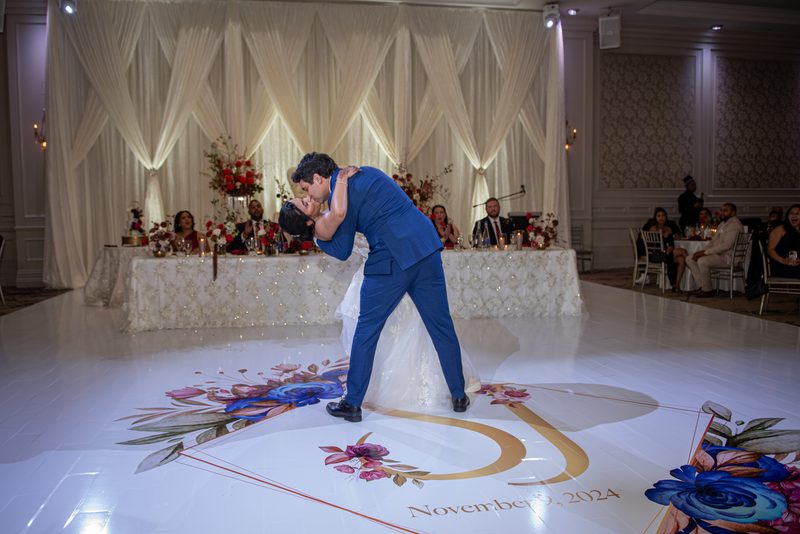 A bride and groom dance intimately on a floral-decorated floor, the groom kissing her forehead as guests watch.