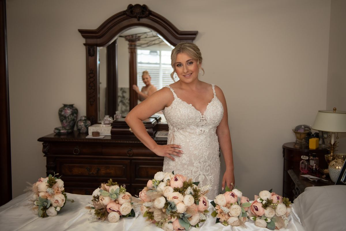 A bride poses confidently in a bedroom, smiling with one hand on her hip and surrounded by floral bouquets.