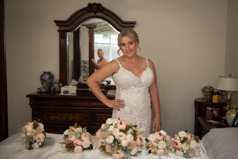 A bride poses confidently in a bedroom, smiling with one hand on her hip and surrounded by floral bouquets.