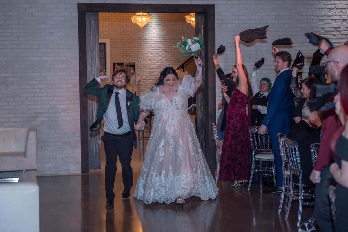 A smiling bride and groom walk through a doorway into a reception where guests toss hats in the air.