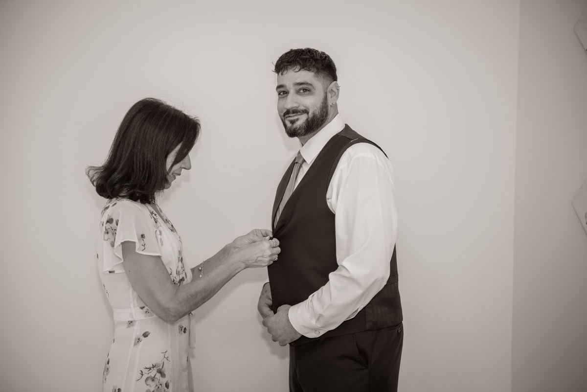 A woman buttons a vest on a smiling man in a tender pre-ceremony moment, illuminated by soft light against a plain wall.