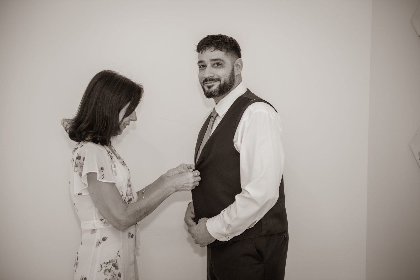 A woman buttons a vest on a smiling man in a tender pre-ceremony moment, illuminated by soft light against a plain wall.