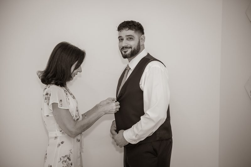 A woman buttons a vest on a smiling man in a tender pre-ceremony moment, illuminated by soft light against a plain wall.