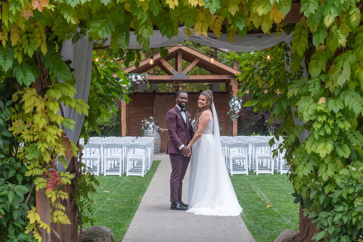 A newlywed couple stands hand-in-hand under a wooden arbor adorned with flowers and string lights, smiling at the camera.