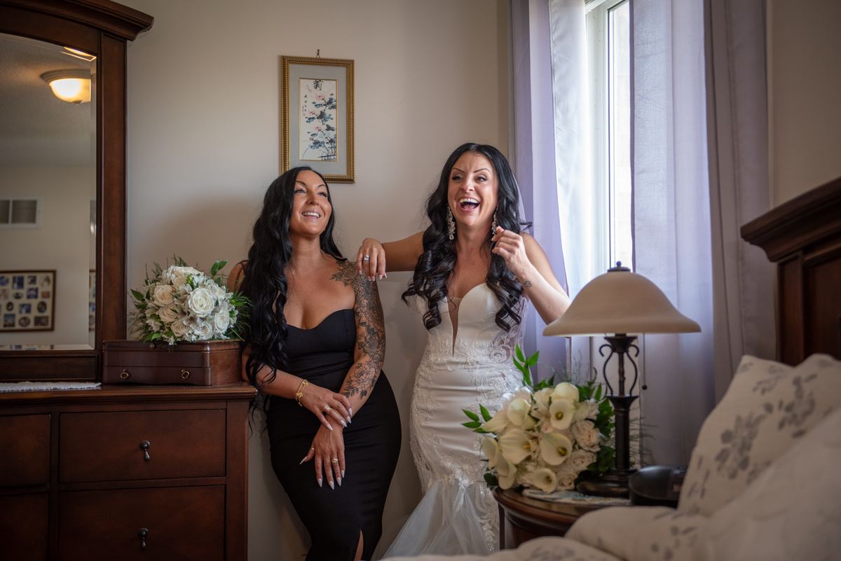 Two women in a bedroom share a joyful moment, one laughing in a wedding gown while the other smiles.